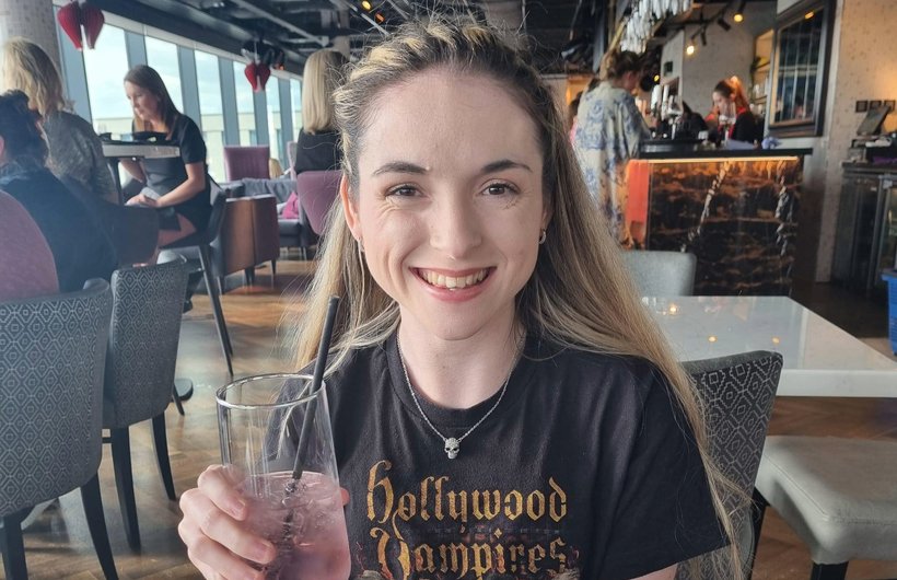 Caitlyn in a bar, smiling at the camera. She is holding a drink and wearing a Hollywood Vampires band t shirt.