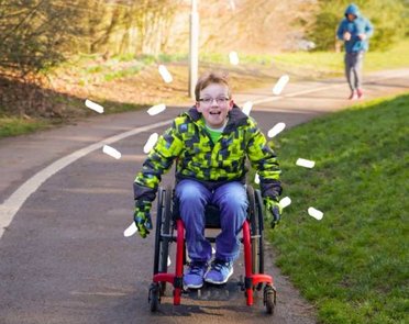A young wheelchair user rolls pushing himself down a park path