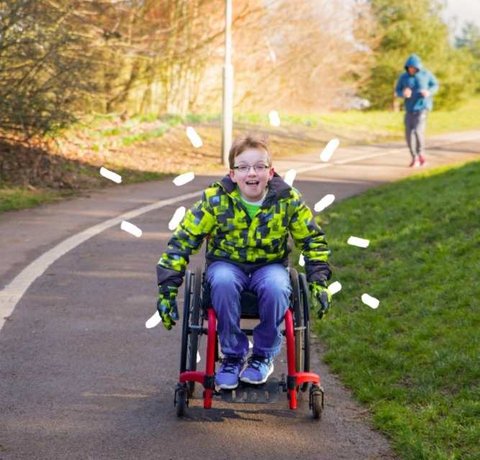 A young wheelchair user rolls pushing himself down a park path