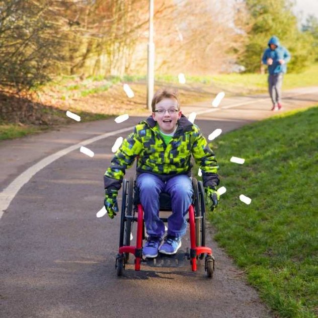 A young wheelchair user rolls pushing himself down a park path