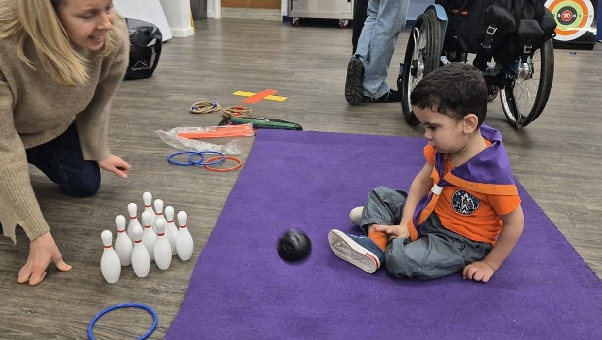 A young scout wearing an orange t shirt throws a small ball at some skittles. There is a wheelchair in the background