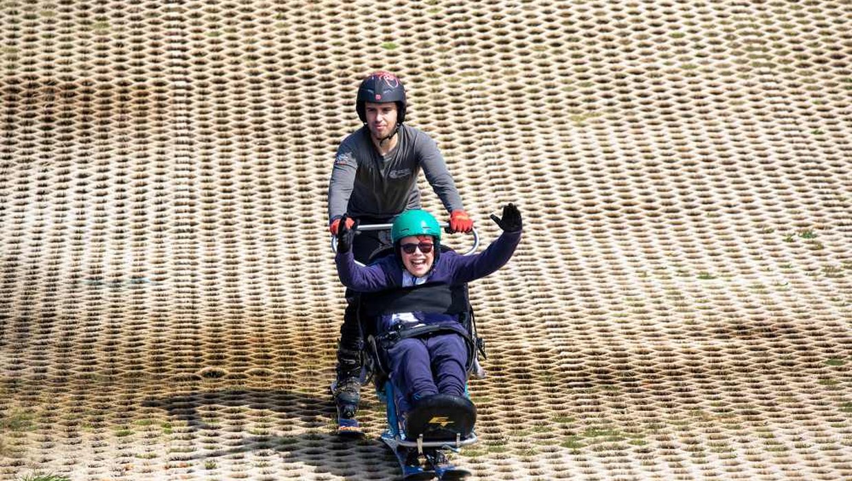 A young wheelchair user is pushed down a dry ski slope in adapted skis
