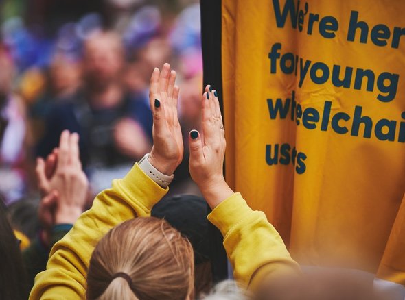 People clapping on the Whizz Kidz cheer point next to a Whizz Kidz flag which reads "we're here for young wheelchair users"