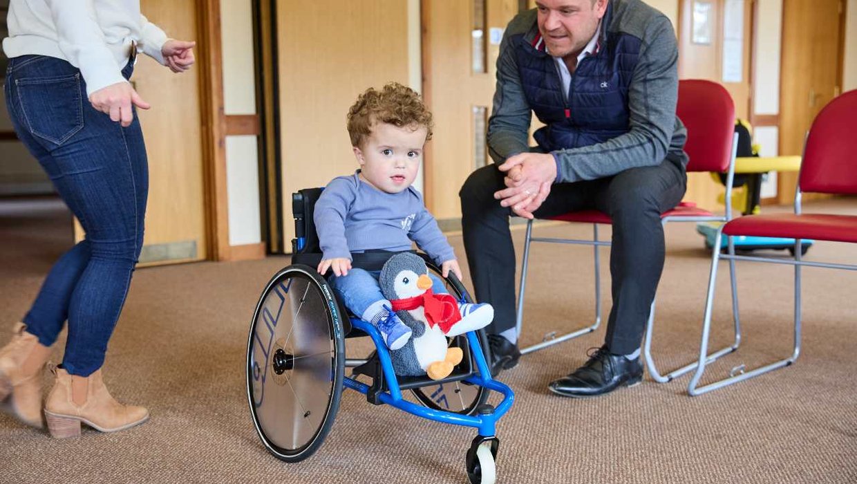 Jake in his wheelchair looking at the camera. He has his toy penguin on the footplate. He is wearing blue Adidas trainers.
