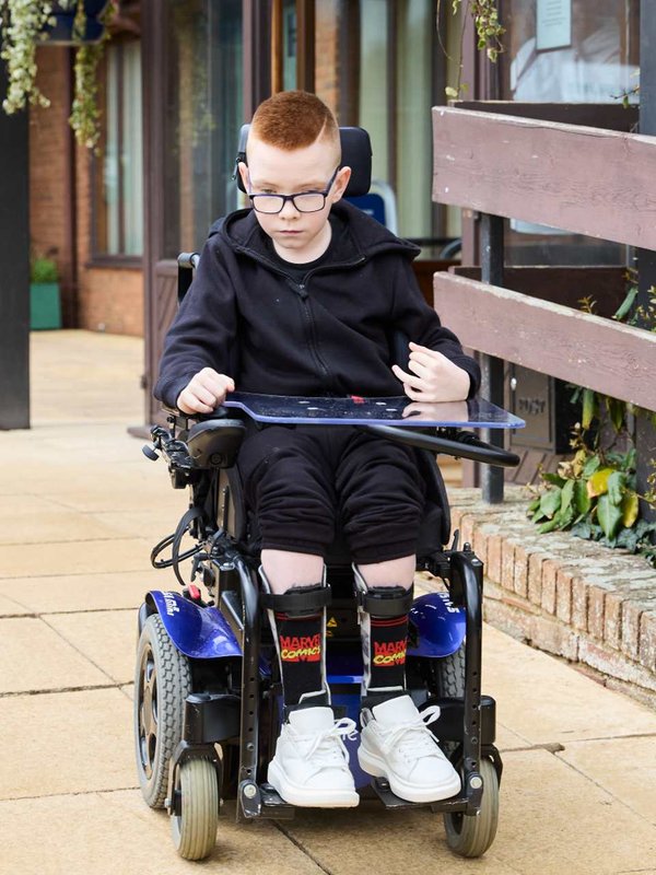 Logan outside in his power chair, using his joystick to steer the chair. He's looking down and concentrating.