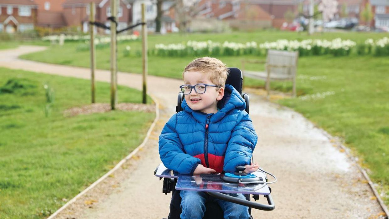 Ethan is wheeling forward in his powered chair. He is wearing a blue puffer jacket and going down a pathway through a park