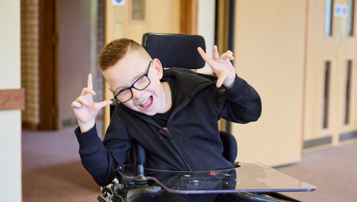 Logan in his wheelchair, posing for the camera. He's holding his hands up and pointing with his thumbs and fingers.