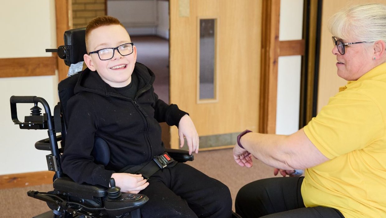 Logan in his wheelchair, smiling for the camera. Sarah Wallace is in front of him, wearing a yellow polo shirt, adjusting his chair.
