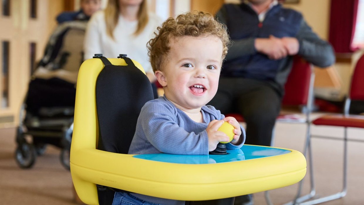 Jake in a powered mobility chair with a yellow tray. He is smiling at the camera and had his hand on the joystick controlling the chair.