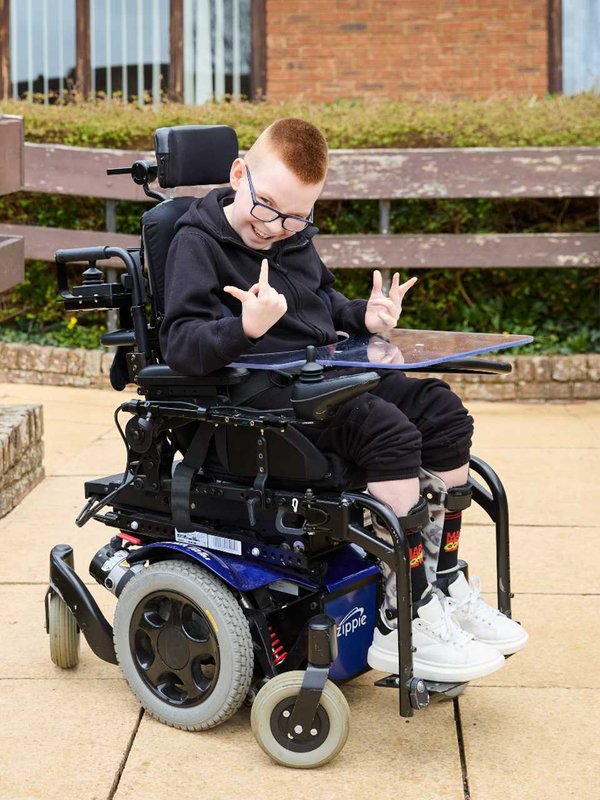 Logan in his wheelchair, posing for the camera. He's holding his hands up and pointing with his thumbs and fingers and smiling. He's outside.