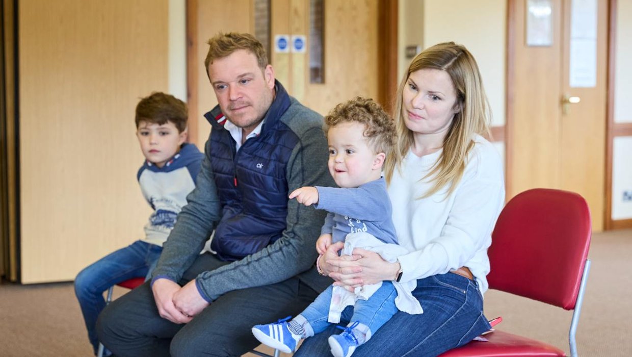 Jake with his family, his brother Jamie, Dad, and Mum, with Jake sitting on his lap on chairs in the Billingshurst clinic.