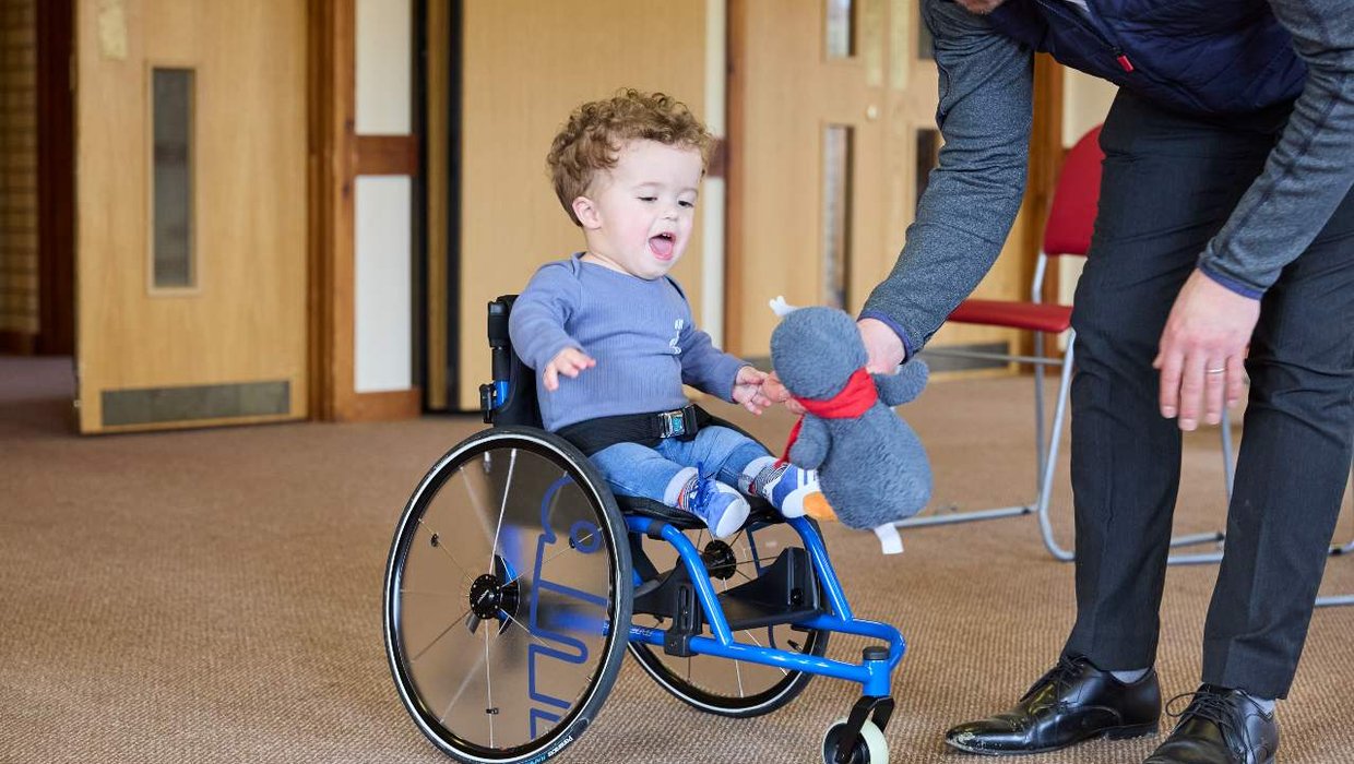 Jake in his wheelchair being given his toy penguin by his Dad.