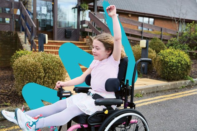 Amber, wearing a pink dress and trainers, whizzes along in her powered chair. Her left arm is raised in the air and the wind is running through her hair.