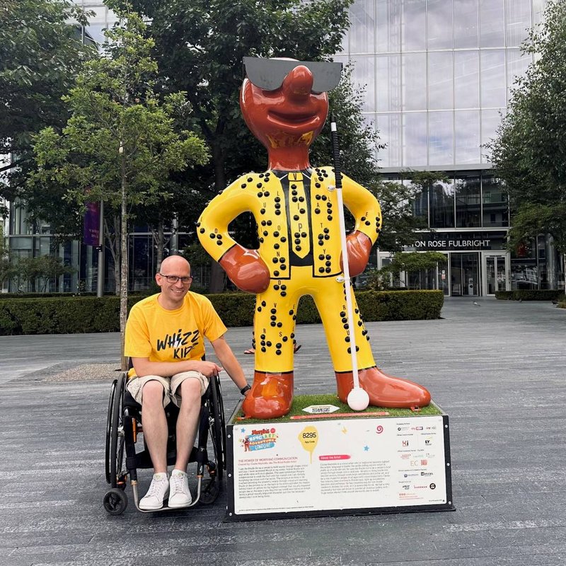 Stephen sits next to a large braille Morph statue. He is wearing a yellow Whizz Kidz t-shirt.