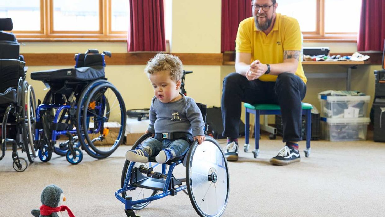 Jake practises pushing by aiming at his toy penguin Quack Quack as Mobility Engineer John Bearman looks on Jake in his wheelchair pushing towards his toy penguin as an engineer looks on.