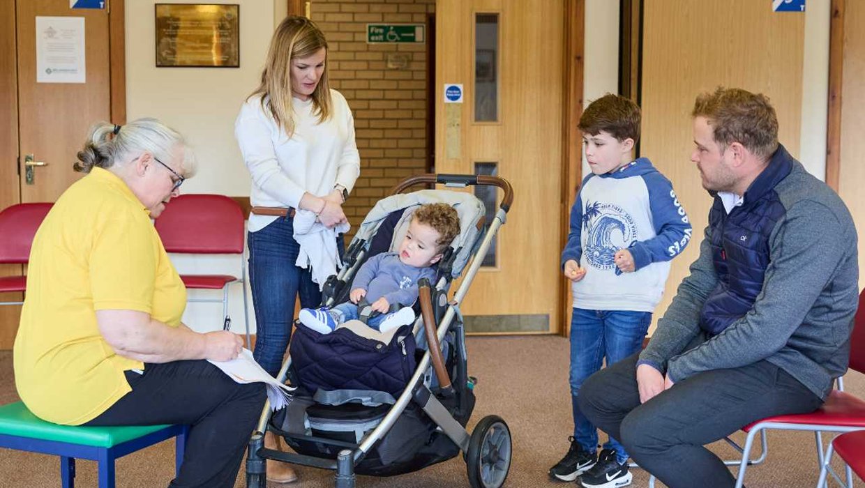 Jake arrives in our clinic with his mum Liz, big brother Jamie and dad, Matt Jake in a buggy with his family in a whizz Kidz clinic. Sarah Wallace is sitting on a stool with a clipboard, facing Jake. Jake's mother, older brother and father surround him.