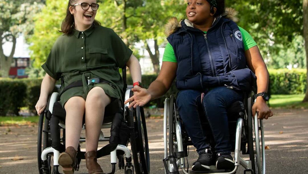 Two members of Whizz Kidz's Kidz Board in their wheelchairs, rolling through a park.