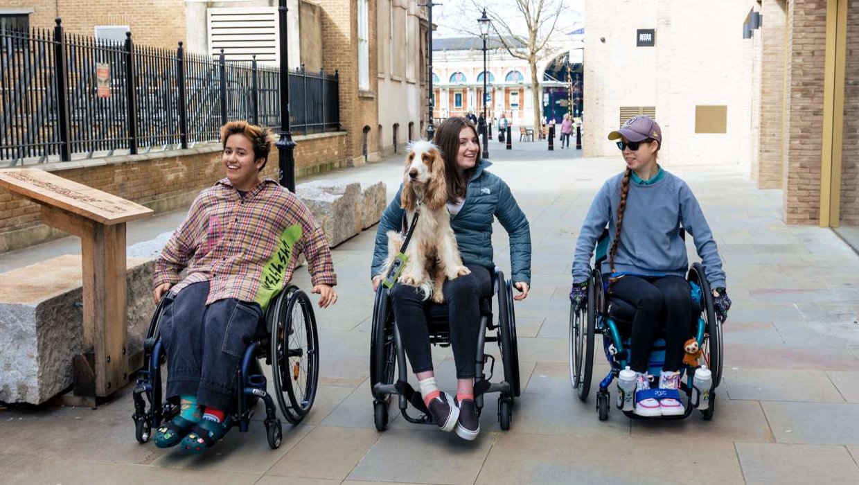 Three young wheelchair users wheel down a pedestrianised street. A super cute dog sits on the lap of the person in the middle.