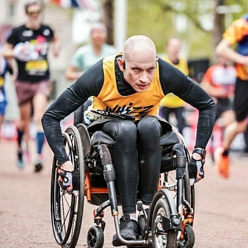 Stephen wheels along The Mall during the London Marathon. He is wearing a yellow Whizz Kidz running vest. There are runners in soft focus behind him and a Union Jack flag in the background.